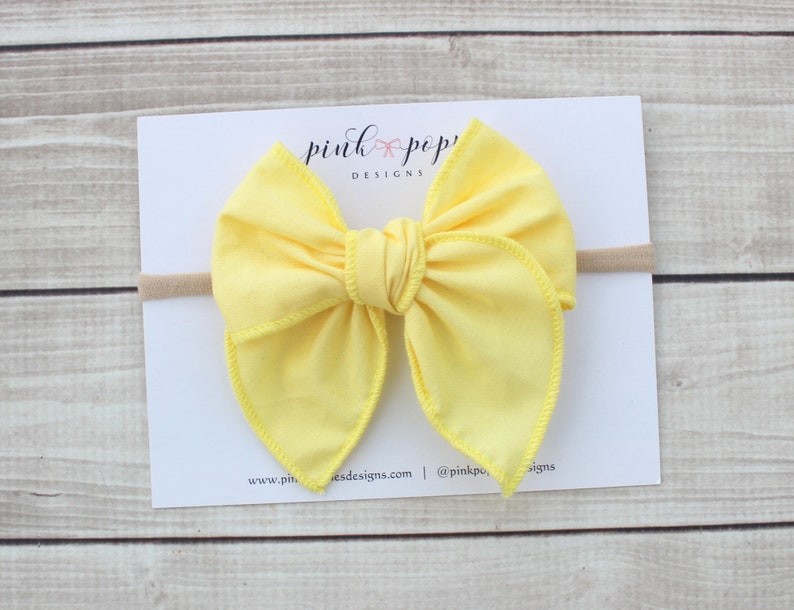 May include: A yellow fabric bow headband with a nude elastic band. The bow is tied in a knot in the center and has a slightly pointed shape. The headband is on a white card with the text "pink poppy designs" printed on it.