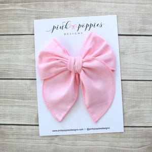 May include: A large, pink fabric bow with a slightly pointed shape. The bow is on a white card with the text "pink poppies designs" and a website address.
