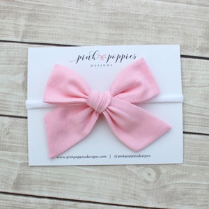 May include: A pink fabric bow headband with a white elastic band. The headband is on a white card with the text "pink poppies designs" and the website address "www.pinkpoppiesdesigns.com" and the social media handle "@pinkpoppiesdesigns".
