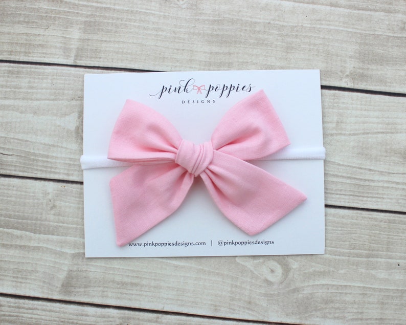 May include: A pink fabric bow headband with a white elastic band. The headband is on a white card with the text "pink poppies designs" and the website address "www.pinkpoppiesdesigns.com" and the social media handle "@pinkpoppiesdesigns".