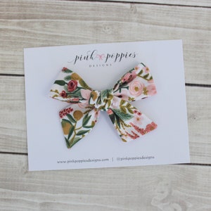 K&ouml;nnte beinhalten: Eine Haarschleife mit floralem Muster in Rosa, Wei&szlig; und Gr&uuml;n. Die Schleife befindet sich auf einer wei&szlig;en Karte mit dem Text "pink poppies designs" und einer Website-Adresse.