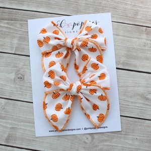 May include: Two white fabric hair bows with an orange pumpkin pattern. The bows are on a white card with the text "Pink Poppies Designs". The bows have orange stitching around the edges.