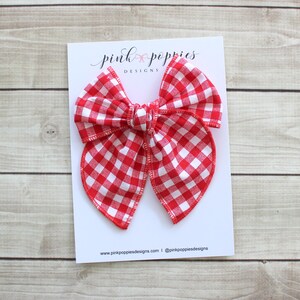 Puede incluir: Lazo de vichy rojo y blanco sobre un fondo blanco. El lazo está atado en un nudo y tiene un borde rizado. El texto "pink poppies designs" está impreso en el fondo blanco.