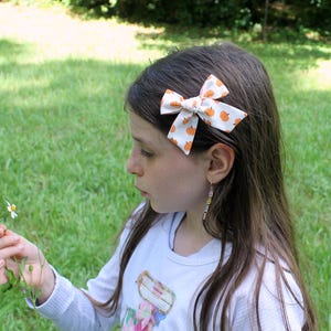 May include: A white hair bow with a pattern of orange pumpkins is clipped into a person's dark hair. The bow is a classic bow shape. The person is wearing a white long-sleeved shirt and holding a small flower.