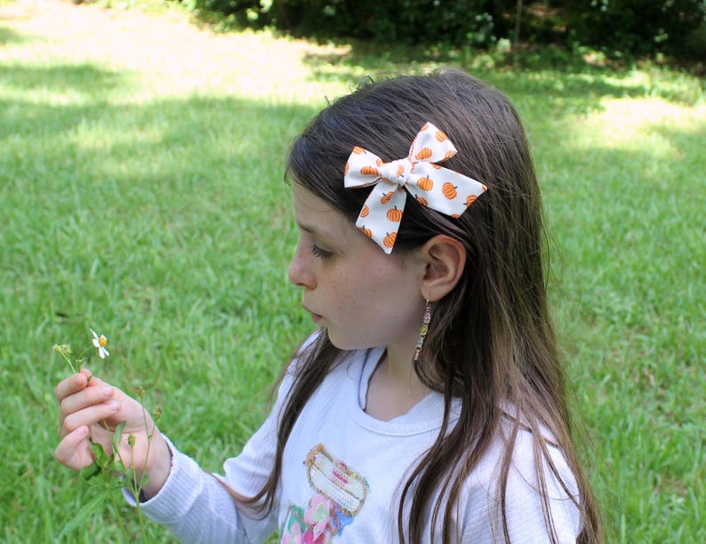 May include: A white hair bow with a pattern of orange pumpkins is clipped into a person's dark hair. The bow is a classic bow shape. The person is wearing a white long-sleeved shirt and holding a small flower.