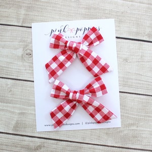May include: Three red and white gingham hair bows on a white card with the text "pink poppers designs" and a website address.