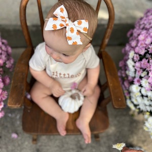 May include: A baby wearing a white onesie with the word "little" printed on it, and a white bow with orange pumpkins. The baby is sitting in a wooden chair, holding a white pumpkin, with purple flowers in the background.