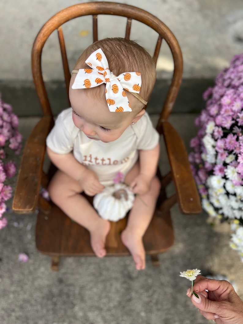 May include: A baby wearing a white onesie with the word "little" printed on it, and a white bow with orange pumpkins. The baby is sitting in a wooden chair, holding a white pumpkin, with purple flowers in the background.