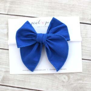 May include: A royal blue fabric bow headband. The bow is tied in a large, loose knot with two long, flowing tails. The headband is white.