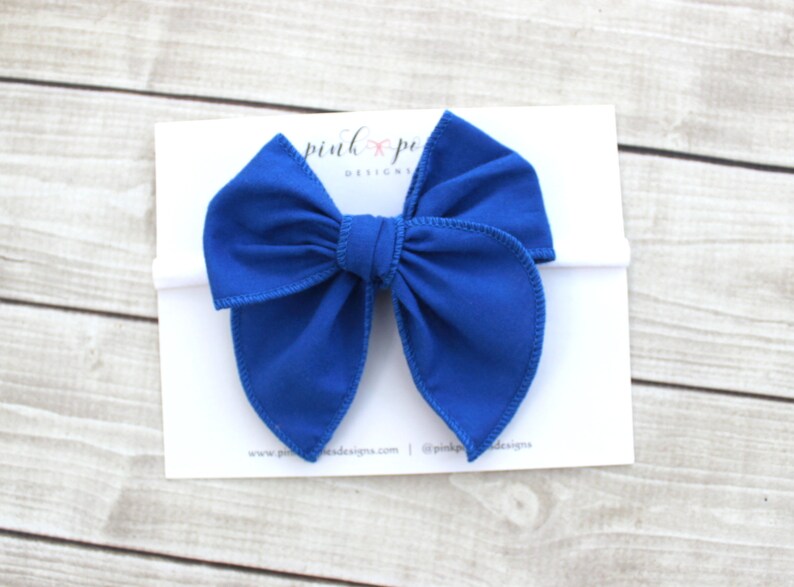 May include: A royal blue fabric bow headband. The bow is tied in a large, loose knot with two long, flowing tails. The headband is white.