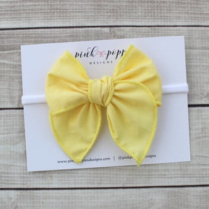 May include: A yellow fabric bow headband with a white elastic band. The headband is on a white card with the text "pink poppy designs" printed on it.