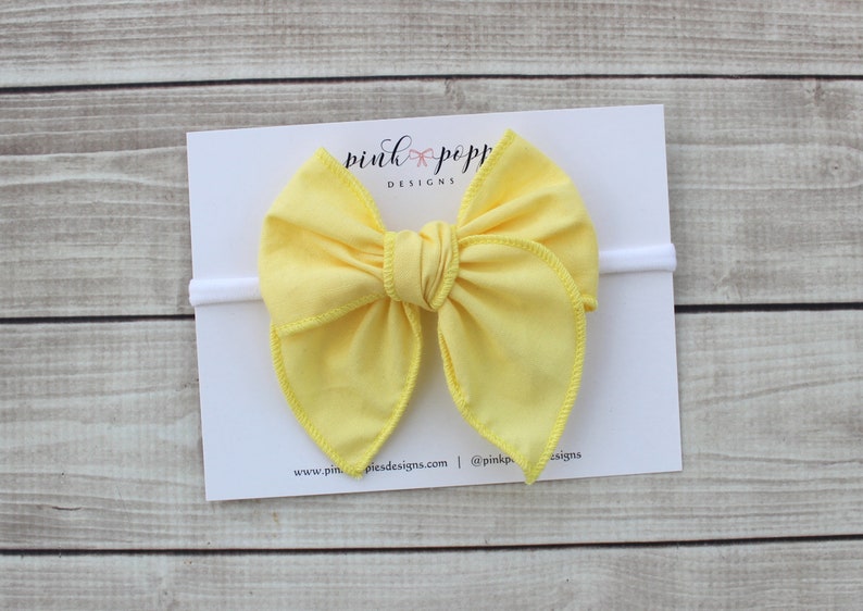 May include: A yellow fabric bow headband with a white elastic band. The headband is on a white card with the text "pink poppy designs" printed on it.