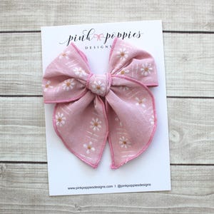 Op de afbeelding: Een grote roze stoffen strik met een madeliefjespatroon en witte accenten. De strik ligt op een witte kaart met de tekst "pink poppies DESIGNS" en website-informatie. Waarschijnlijk een haaraccessoire.