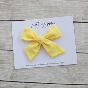 May include: A bright yellow fabric bow with a center knot. The bow is on a white card with the text "pink poppies designs" and website information.