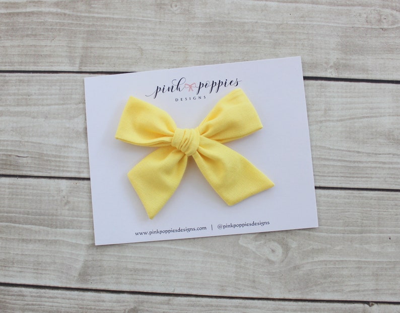 May include: A bright yellow fabric bow with a center knot. The bow is on a white card with the text "pink poppies designs" and website information.