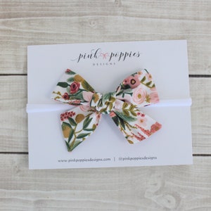 K&ouml;nnte beinhalten: Ein wei&szlig;es Stirnband mit einer Schleife im Blumenmuster. Die Schleife besteht aus einem wei&szlig;en Stoff mit rosa, roten und gr&uuml;nen Blumen. Das Stirnband ist zu einer Schleife gebunden und bereit zum Tragen.