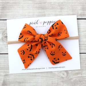 May include: An orange Halloween hair bow featuring a repeating black jack-o'-lantern face pattern. The bow is attached to a beige headband and rests on a white card with the text "pink poppies designs". The background is a light wood grain.