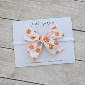 May include: A white baby headband with a bow featuring a pattern of orange pumpkins. The bow is centered on a white card with the text "pink poppies designs" in cursive. The background is a light wood surface.