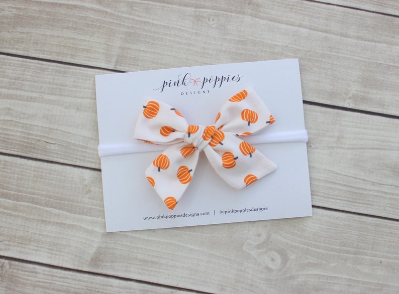 May include: A white baby headband with a bow featuring a pattern of orange pumpkins. The bow is centered on a white card with the text "pink poppies designs" in cursive. The background is a light wood surface.