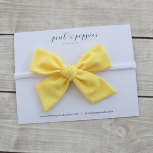 May include: A yellow fabric bow headband on a white background. The headband is made of a soft, stretchy material and is perfect for babies and toddlers.
