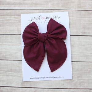 May include: A large, burgundy fabric bow on a white card with the text "pink poppies DESIGNS". The bow is centered on the card, with a simple, elegant design. The card also includes the website address and social media handle.