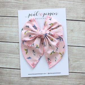 Op de afbeelding: Een grote roze haarstrik met een bloemenpatroon met kleine blauwe, gele en witte bloemen. De strik is gecentreerd op een witte kaart met de tekst "pink poppies designs" in cursief. De strik is ongeveer 15 cm breed.