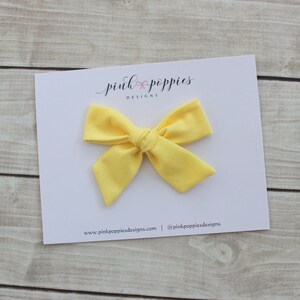 May include: A bright yellow fabric bow with a simple knot in the center. The bow is on a white card with the text "pink poppies designs" and website information.