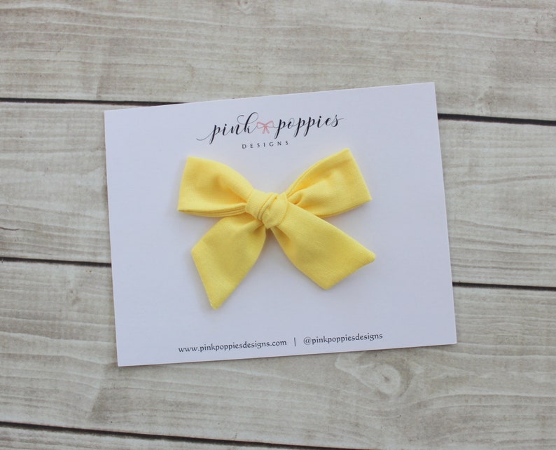 May include: A bright yellow fabric bow with a simple knot in the center. The bow is on a white card with the text "pink poppies designs" and website information.