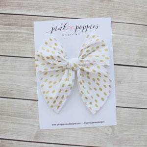 May include: White fabric bow with gold polka dots. The bow is on a white card with the text "pink poppies designs" and the website address. The bow is a hair accessory.