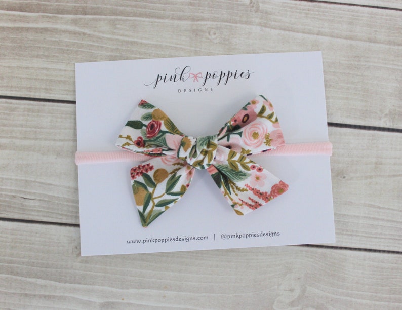 K&ouml;nnte beinhalten: Ein rosa Haarband mit einem floralen Muster in Rosa. Die Schleife ist in einem Knoten gebunden und hat einen wei&szlig;en Hintergrund mit rosa, roten und gr&uuml;nen Blumen. Der Text "pink poppies designs" ist auf einer wei&szlig;en Karte hinter der Schleife gedruckt.