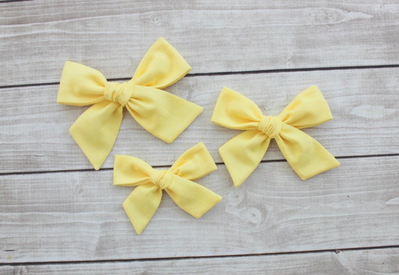 May include: Three yellow fabric hair bows of different sizes. The bows are tied in a knot in the center and have a soft, fluffy texture.