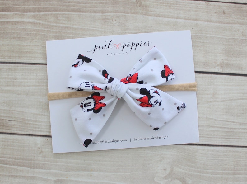 May include: A white headband with a large bow featuring a Minnie Mouse print. The bow is made of white fabric with gray polka dots and red accents. The headband is made of a nude elastic material.
