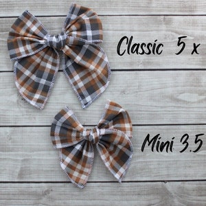 May include: Two plaid fabric hair bows in brown, gray, and white. The larger bow is labeled "Classic 5 x 5.5"" and the smaller bow is labeled "Mini 3.5 x 4"". The bows are on a light wood grain background.