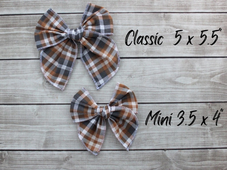 May include: Two plaid fabric hair bows in brown, gray, and white. The larger bow is labeled "Classic 5 x 5.5"" and the smaller bow is labeled "Mini 3.5 x 4"". The bows are on a light wood grain background.
