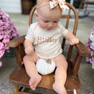 May include: A baby wearing a cream-colored onesie with the text "little pumpkin" in brown lettering. The baby is seated in a wooden rocking chair and wearing a headband with a pumpkin-patterned bow. A white pumpkin sits in the baby's lap.