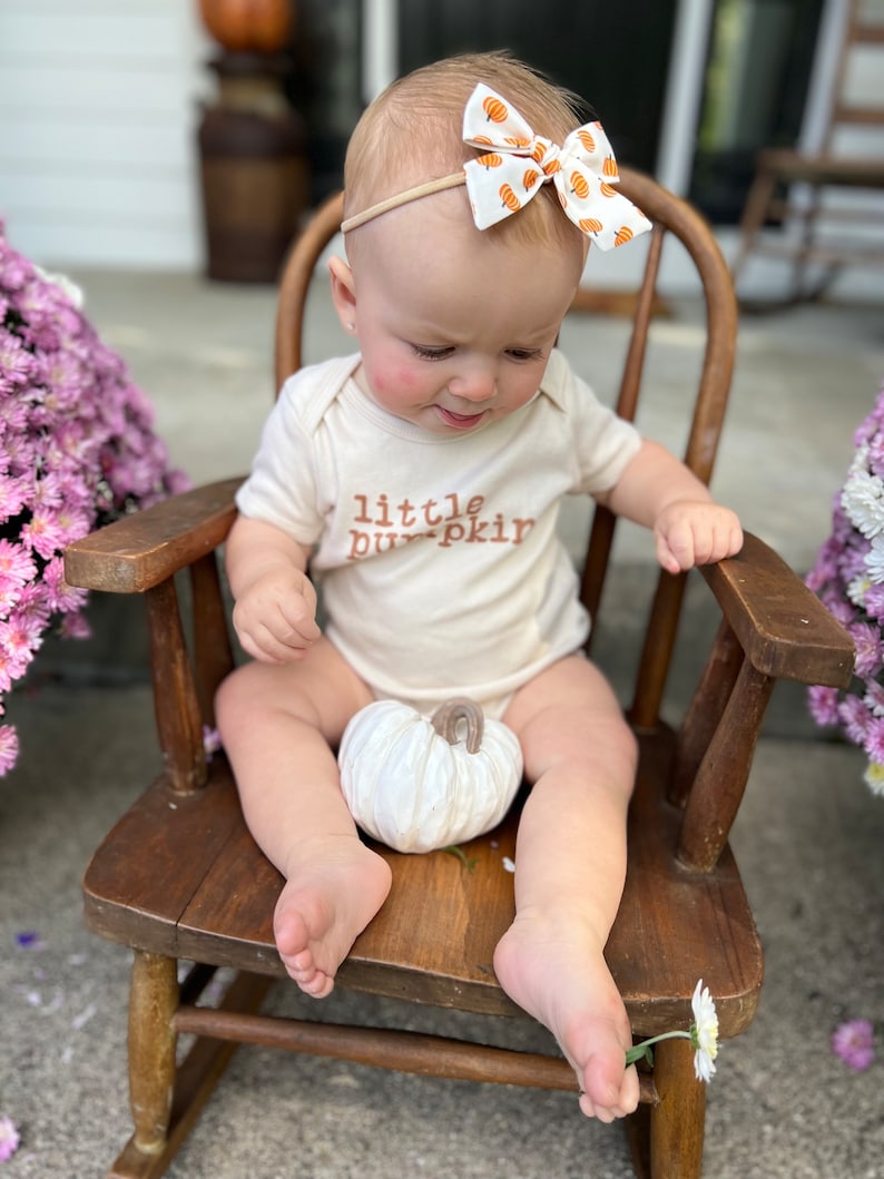 May include: A baby wearing a cream-colored onesie with the text "little pumpkin" in brown lettering. The baby is seated in a wooden rocking chair and wearing a headband with a pumpkin-patterned bow. A white pumpkin sits in the baby's lap.