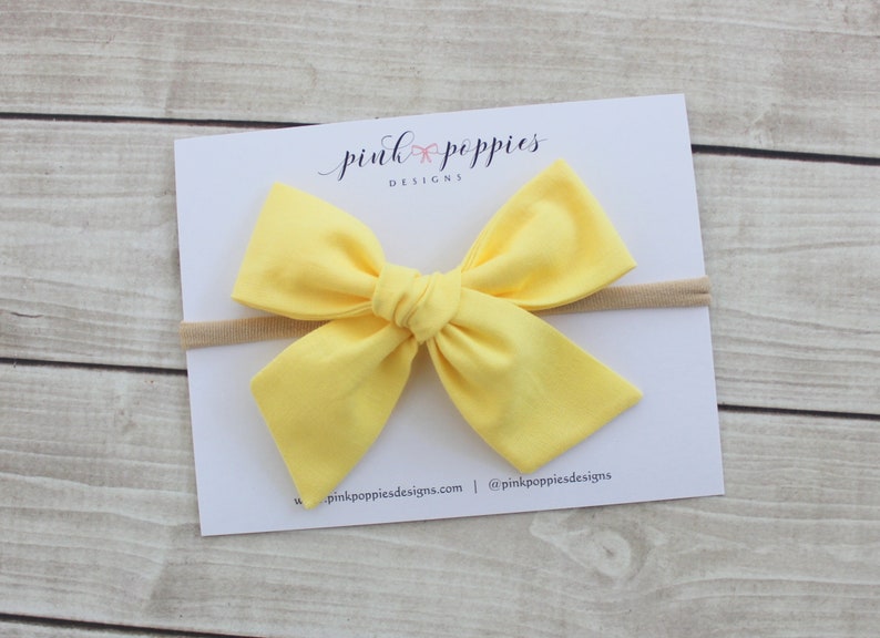 May include: A yellow fabric bow headband with a nude elastic band. The headband is on a white card with the text "pink poppies designs" in black.