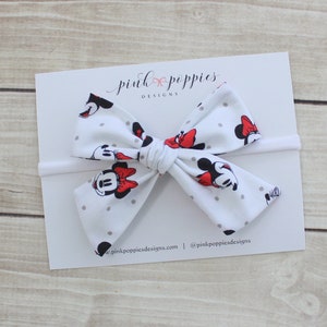 May include: White headband with a large bow featuring a Minnie Mouse print with red polka dots. The bow is tied in a knot and the headband is made of elastic.