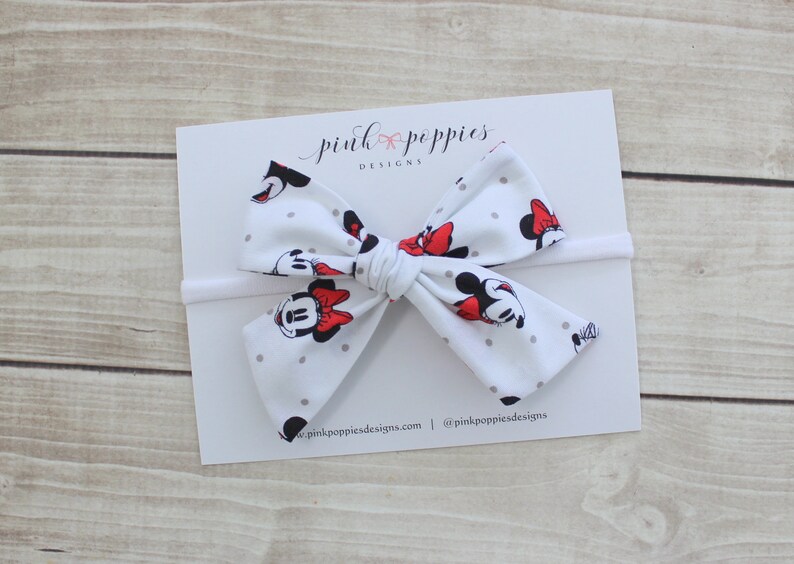 May include: White headband with a large bow featuring a Minnie Mouse print with red polka dots. The bow is tied in a knot and the headband is made of elastic.