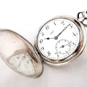 May include: Open silver pocket watch with a white face, black hands, and black Arabic numerals. The watch has a small seconds dial and a gold-colored crown. The watch is open, revealing the inside of the lid.