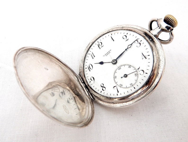 May include: Open silver pocket watch with a white face, black hands, and black Arabic numerals. The watch has a small seconds dial and a gold-colored crown. The watch is open, revealing the inside of the lid.