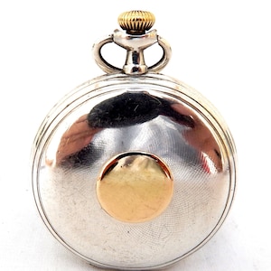 May include: A silver pocket watch with a gold-colored crown and a gold-colored circular accent on the face. The watch has a round shape and a textured surface. The watch is a classic timepiece.
