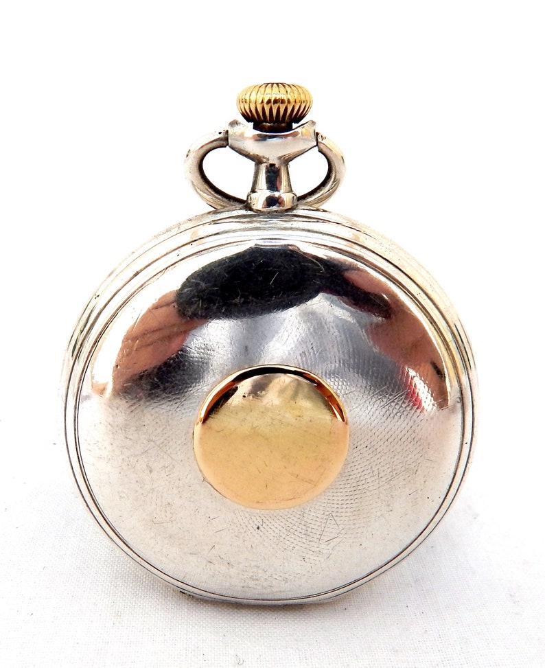 May include: A silver pocket watch with a gold-colored crown and a gold-colored circular accent on the face. The watch has a round shape and a textured surface. The watch is a classic timepiece.