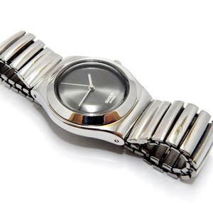 Puede incluir: Un reloj Swatch de cuarzo plateado con una esfera gris oscura y una correa de metal flexible y extensible. El reloj tiene un diseño minimalista con una esfera redonda y agujas sencillas. El reloj se muestra sobre un fondo blanco.