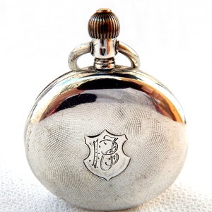 K&ouml;nnte beinhalten: Eine silberne Taschenuhr mit einem gravierten Muster auf der R&uuml;ckseite. Die Uhr hat eine Krone und eine Schlaufe f&uuml;r eine Kette. Das Zifferblatt der Uhr zeigt ein Schild mit den Initialen "JB" in einer eleganten Schrift.