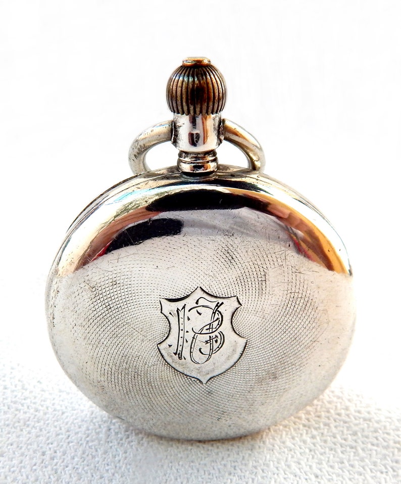 K&ouml;nnte beinhalten: Eine silberne Taschenuhr mit einem gravierten Muster auf der R&uuml;ckseite. Die Uhr hat eine Krone und eine Schlaufe f&uuml;r eine Kette. Das Zifferblatt der Uhr zeigt ein Schild mit den Initialen "JB" in einer eleganten Schrift.
