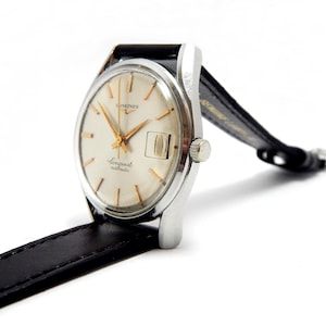 Puede incluir: Un reloj autom&aacute;tico Longines Conquest vintage con caja plateada y esfera color crema. El reloj presenta agujas e indicadores dorados, una ventana de fecha y una correa de cuero negro. Sobre un fondo blanco.