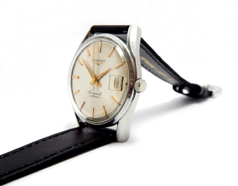 Puede incluir: Un reloj autom&aacute;tico Longines Conquest vintage con caja plateada y esfera color crema. El reloj presenta agujas e indicadores dorados, una ventana de fecha y una correa de cuero negro. Sobre un fondo blanco.