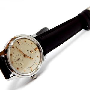 May include: A vintage Omega Automatic wristwatch with a silver case and a cream-colored face. The watch features gold hands and hour markers, a small seconds dial, and a black leather strap. The watch is a classic example of mid-century design.