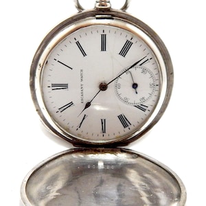 May include: A silver pocket watch with a white face and black Roman numerals. The watch has a small seconds dial and the words "ESCASANY WATCH" are printed on the face. The watch's cover is open, revealing the inside.